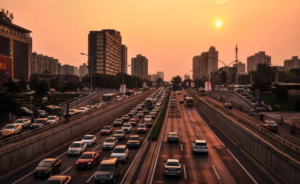 vehicle-in-road-at-golden-hour-210182 A bustling urban highway with heavy traffic under a vibrant sunset sky against a cityscape backdrop.